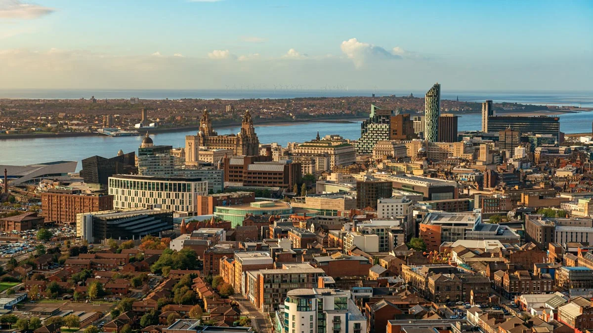 An expansive aerial cityscape view of central Liverpool and the River Mersey, showcasing historic waterfront architecture alongside contemporary residential towers