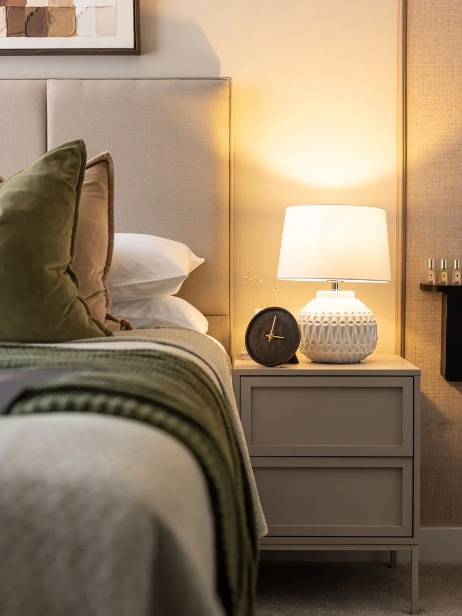 Close-up of a modern bedroom nightstand featuring a white patterned ceramic table lamp, a minimalist black desk clock, and layered green and neutral bedding