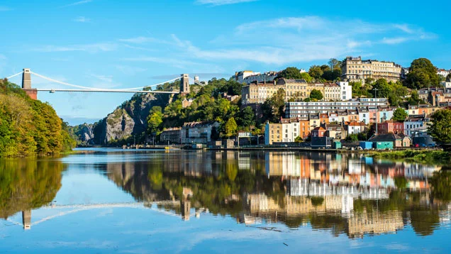 A scenic reflection of the Clifton Suspension Bridge, colourful Georgian terraced houses, and lush green hills on the calm waters of the Avon Gorge in Bristol