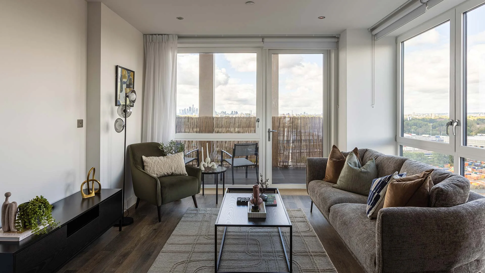 Modern living room with a grey sofa, green armchair, coffee table, and large windows overlooking the city.