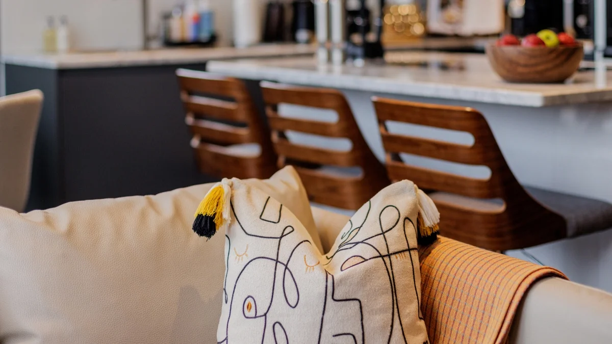 Airbnb living room with decorative cushions and a warm throw, with a modern kitchen in the background.