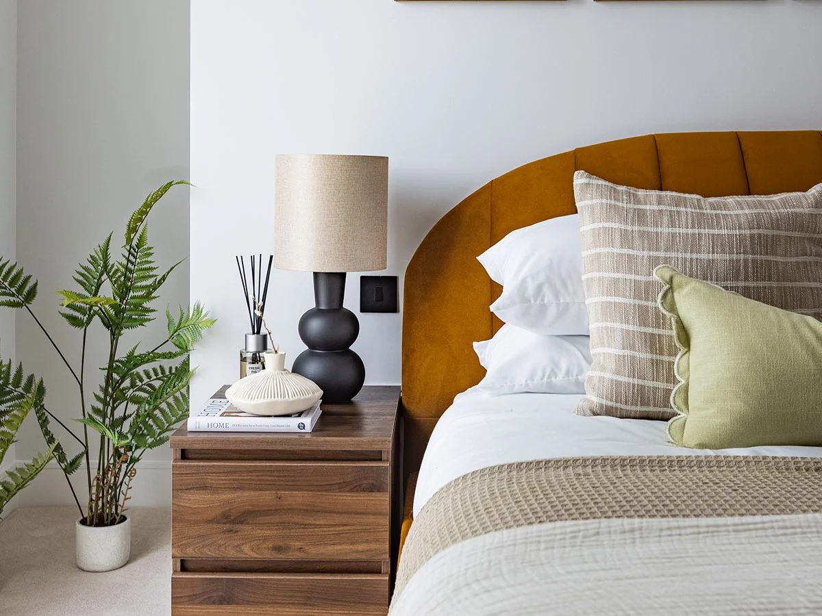 A luxury bedroom corner styled by InStyle Direct at Regents View, showing an ochre velvet headboard, a dark walnut nightstand with a sculpted black table lamp, and a potted indoor fern