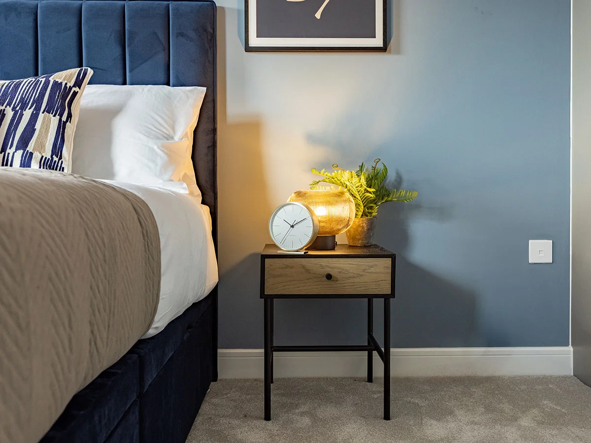 Contemporary bedside table with decorative lighting, clock and plant beside a velvet bed in a professionally furnished London apartment.