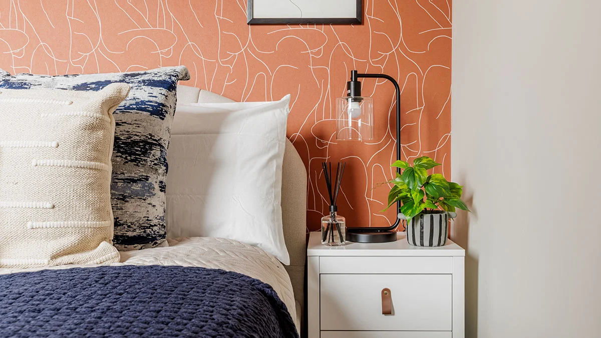 Close-up of a bespoke bedroom design by InStyle Direct showcasing a white nightstand with a glass industrial lamp, a navy textured quilt, and terracotta line-art wallpaper featuring abstract faces in London