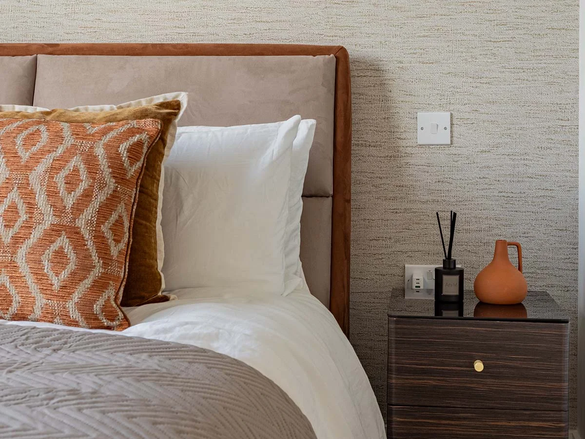 A bespoke bedroom setup by InStyle Direct featuring a plush tan suede headboard, geometric patterned orange accent pillows, a dark wood nightstand, and textured neutral wallpaper in Canary Wharf
