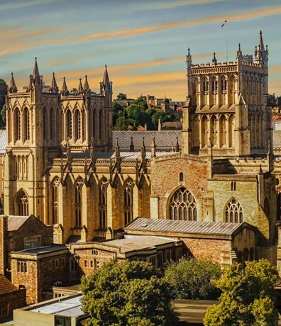 An elevated view of the historic Gothic architecture and ornate stone towers of Bristol Cathedral bathed in warm sunlight under a blue sky