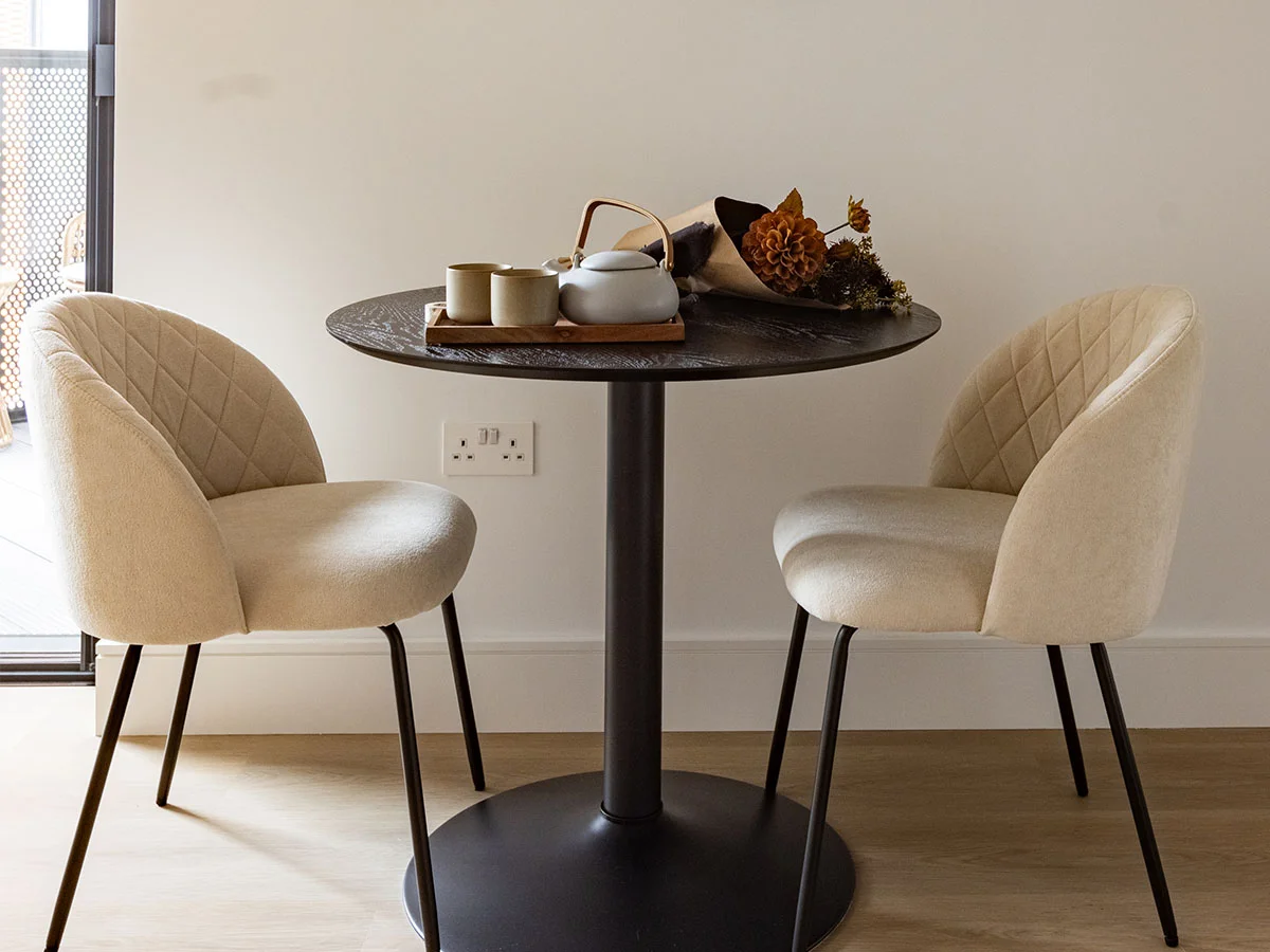 A compact dining nook styled by InStyle Direct at Regents View featuring a black pedestal table with ceramic tea set decor, flanked by cream quilted tub dining chairs