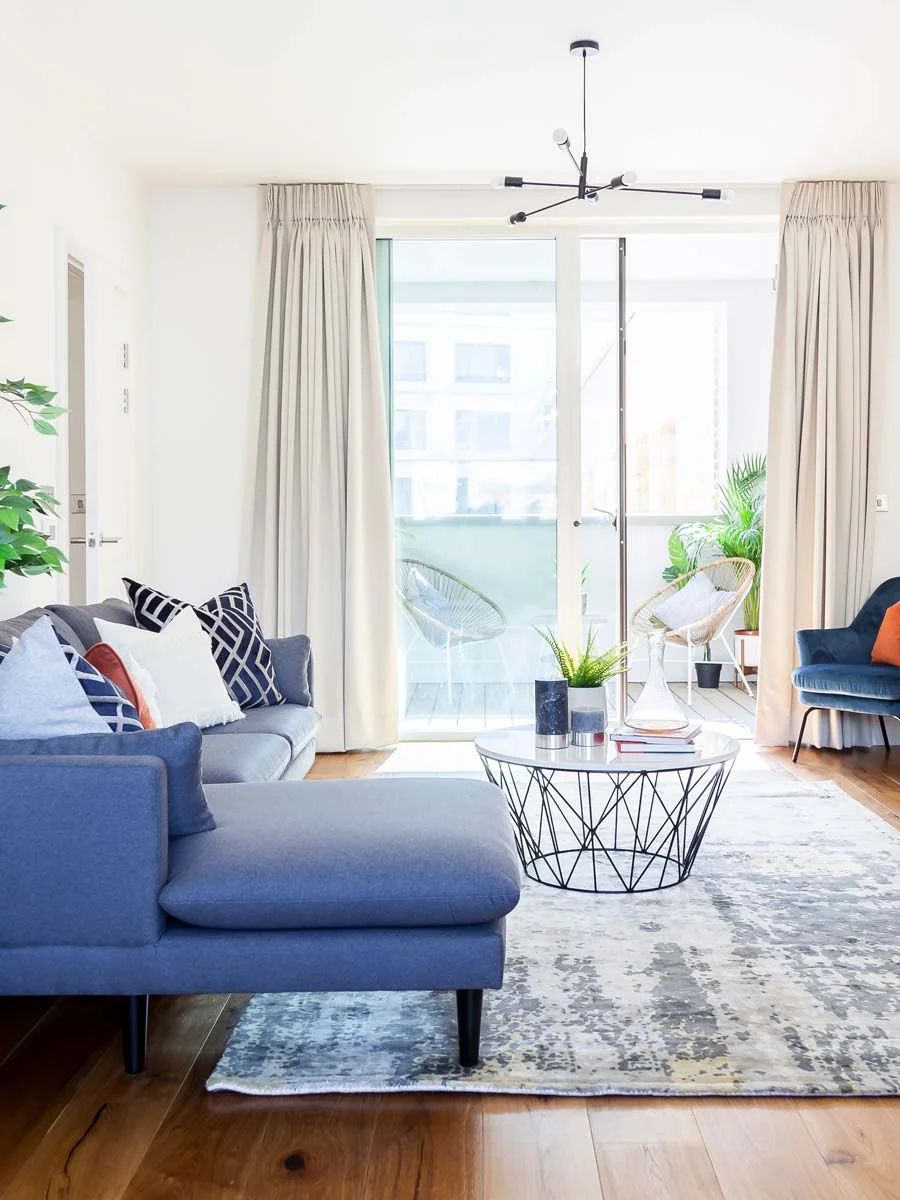 Contemporary living room interior featuring a blue chaise sofa, a round wire-frame coffee table, a patterned rug, and access to a balcony with outdoor seating