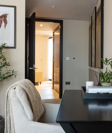 A contemporary home office desk corner styled by InStyle Direct, featuring a sleek black desk with decorative trays, a grey armchair with a textured throw, and a doorway leading into a bright living space