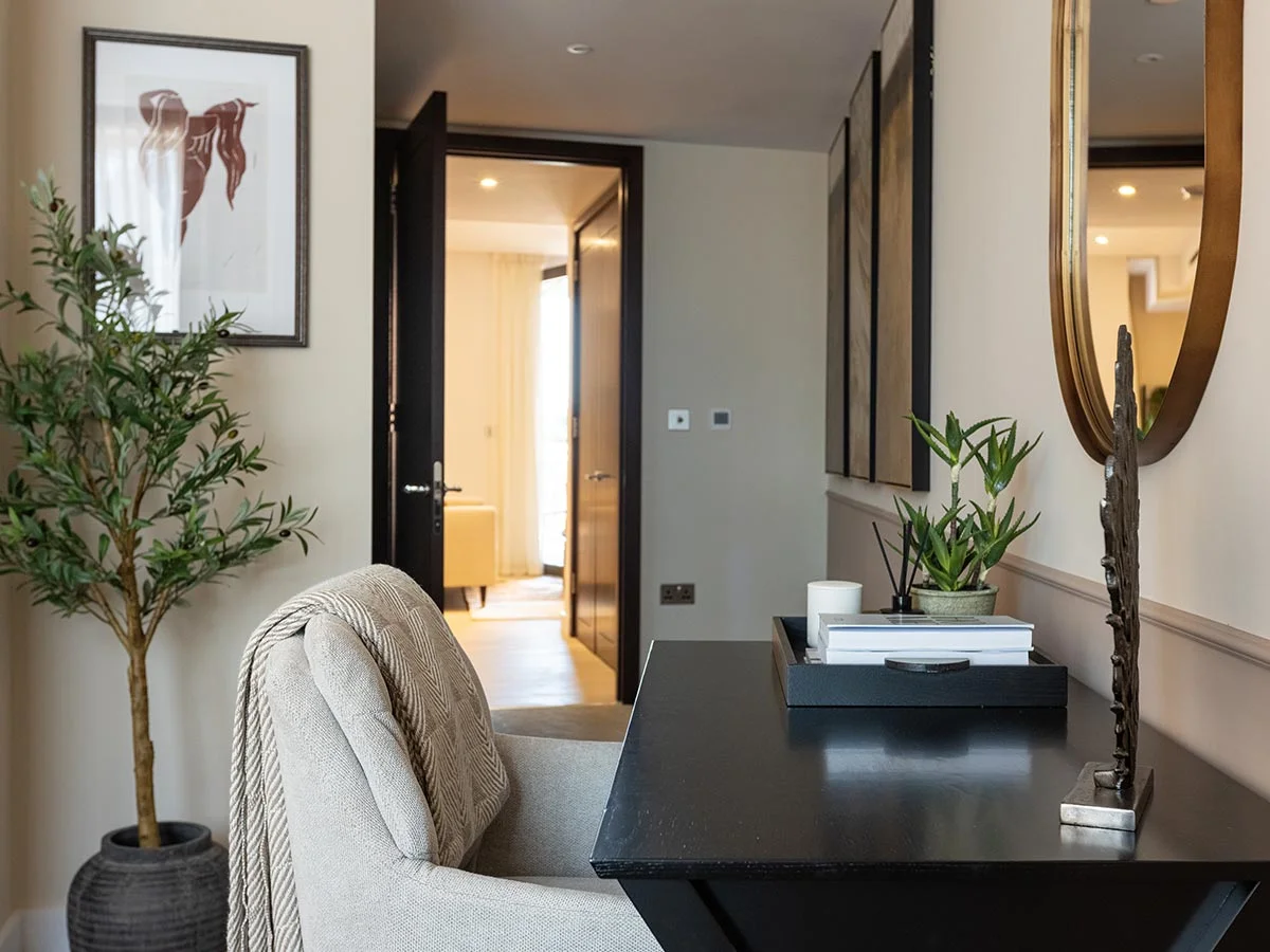 A contemporary hallway workspace styled by InStyle Direct at West End Gate, featuring a sleek black console desk, a light grey upholstered office chair with a textured throw, a golden wall mirror, and a potted olive tree