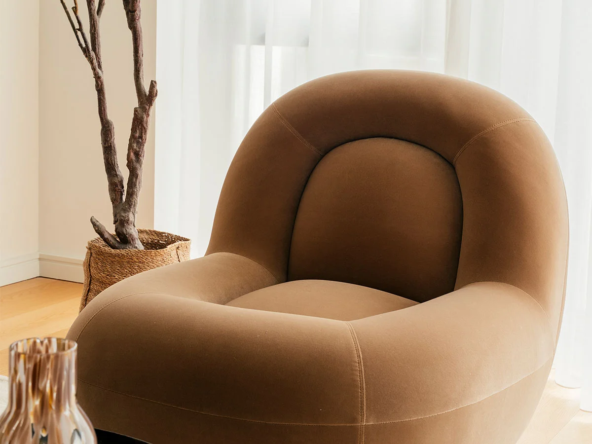 Close-up of a contemporary velvet tub chair in a rich tan color, styled by InStyle Direct next to a potted indoor plant and sheer white floor-to-ceiling drapes in Kensington