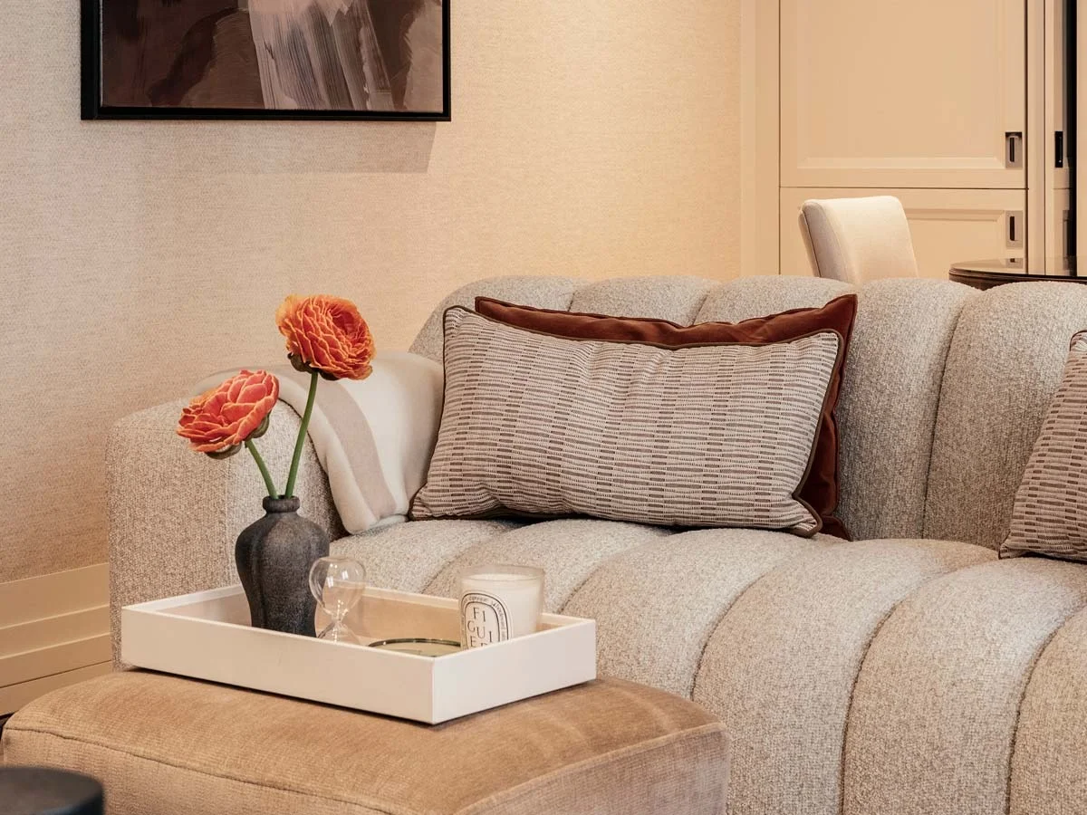 Close-up of a modern textured sofa featuring patterned scatter cushions with dark velvet borders, an upholstered ottoman with a white tray, orange faux flowers in a dark vase, and framed abstract wall art at 60 Curzon, Mayfair