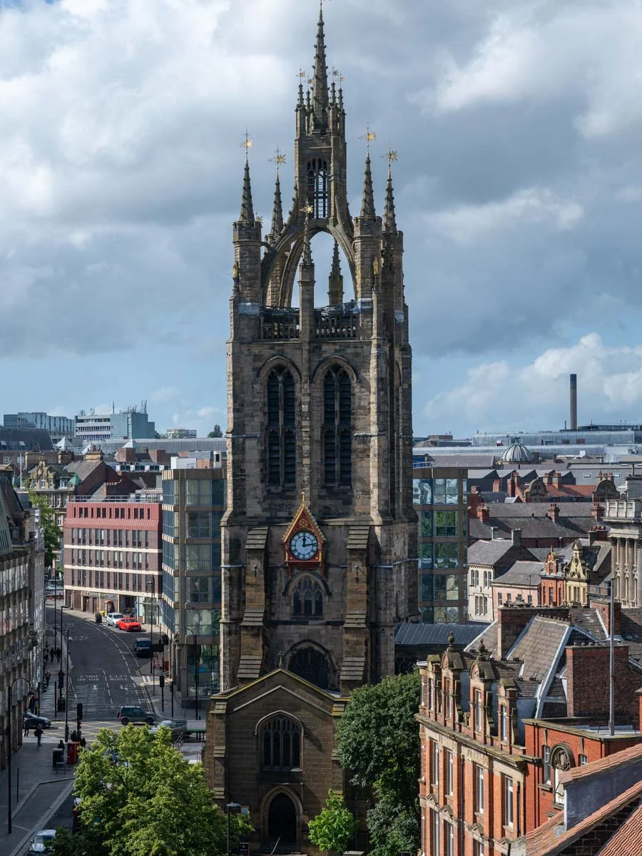 Majestic gothic cathedral tower with ornate spires, a clock face, and surrounding historic urban rooftops under a dramatic cloudy sky