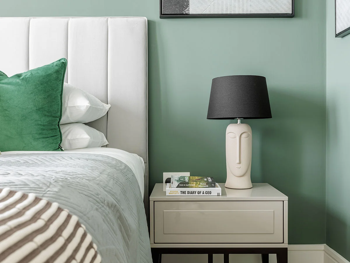 A luxury bedroom corner styled by InStyle Direct with a sage green feature wall, a white panelled headboard with an emerald green velvet cushion, and a unique face-profile ceramic table lamp