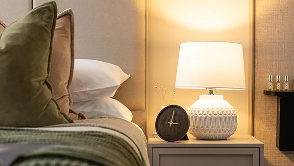 Close-up of a luxury bedroom nightstand staged by InStyle Direct at West End Gate, featuring a white textured geometric table lamp, a round black clock, and plush sage green velvet cushions in Paddington