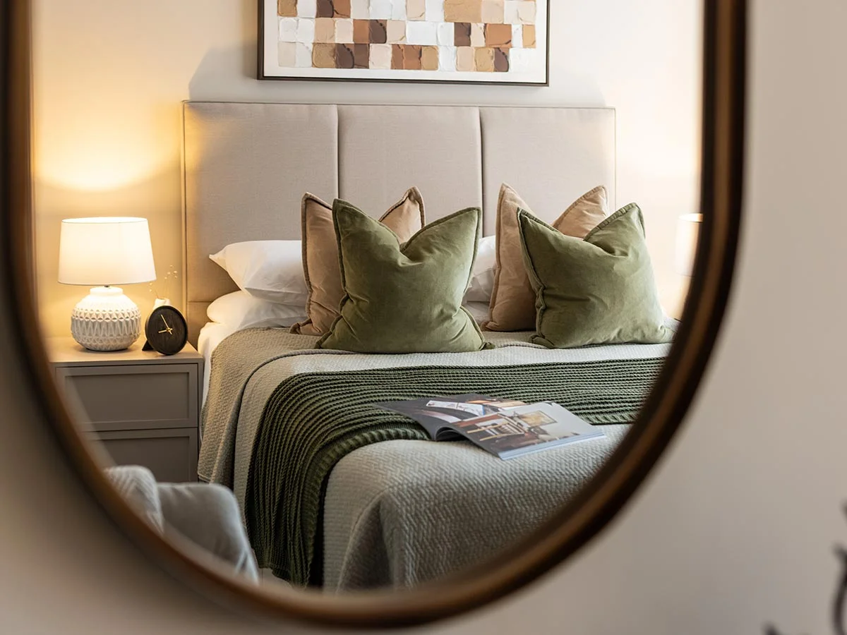 An artistic reflection in an oval mirror showcasing a luxury bedroom furniture set staged by InStyle Direct at West End Gate, featuring a cream headboard, sage green velvet cushions, and an textured abstract painting