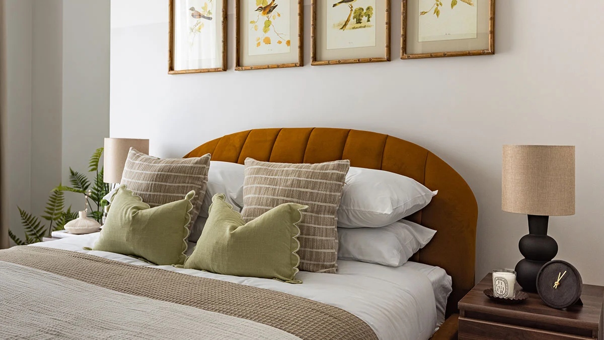 Sophisticated bedroom interior styled with a curved mustard yellow velvet headboard, sage green scatter cushions, neutral bedding, and framed bird illustrations