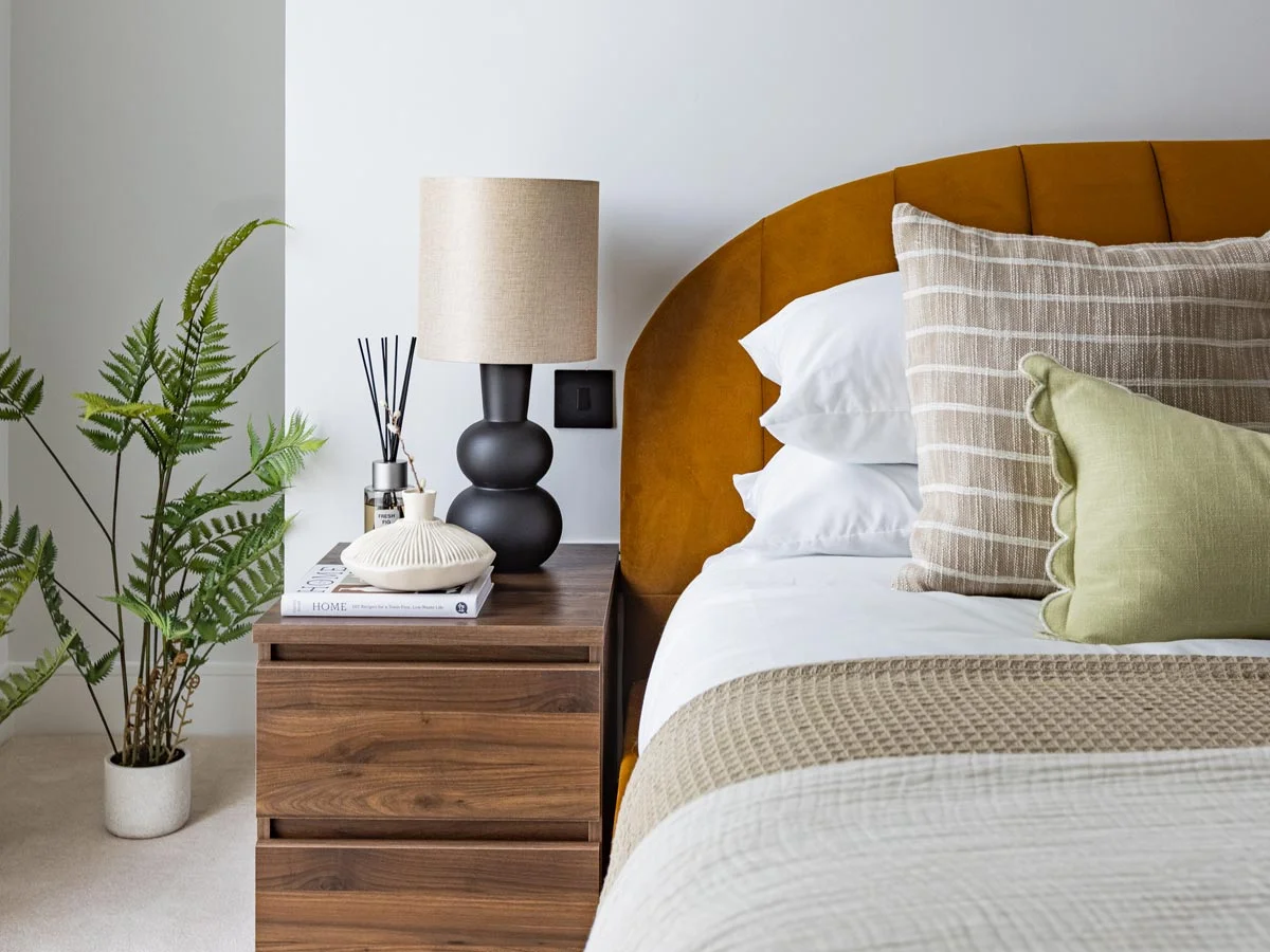 Close-up of bedroom nightstand styling by InStyle Direct at Regents View, featuring a charcoal grey and gold drawer unit, a black bobble table lamp, geometric desk art, and olive green velvet pillows