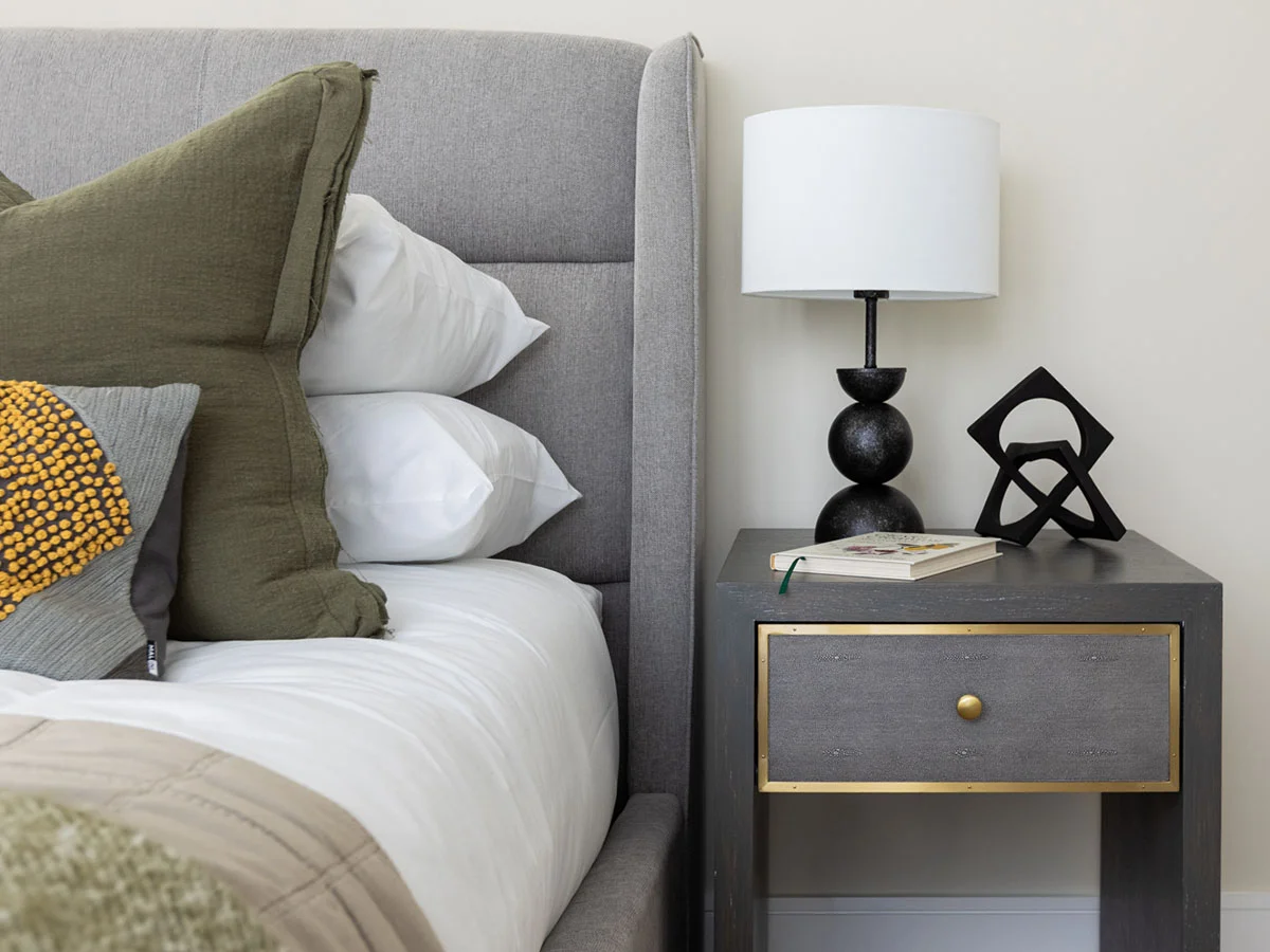 Close-up of bedroom nightstand styling by InStyle Direct at Regents View, featuring a charcoal grey and gold drawer unit, a black bobble table lamp, geometric desk art, and olive green velvet pillows