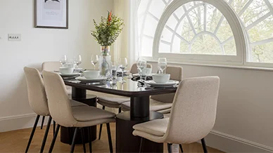 A luxury dining room setup styled by InStyle Direct at Solasta Riverside, Buchanan Wharf, featuring a dark glass-top table, cream upholstered chairs, and a large architectural fanlight window