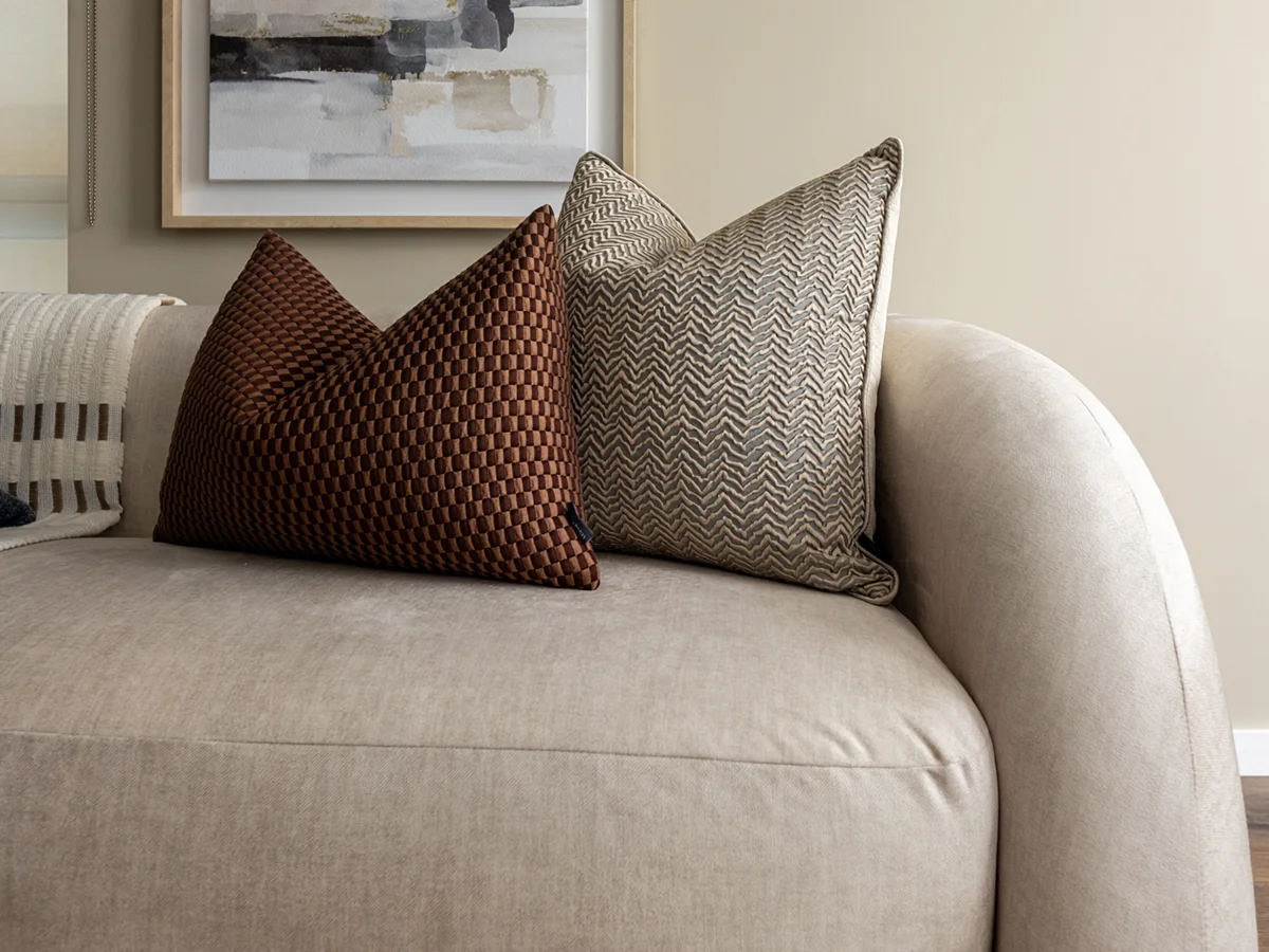 Close-up of high-end sofa styling by InStyle Direct showcasing a neutral fabric sofa adorned with a brown checkered velvet cushion and a grey chevron textured pillow beneath abstract framed art