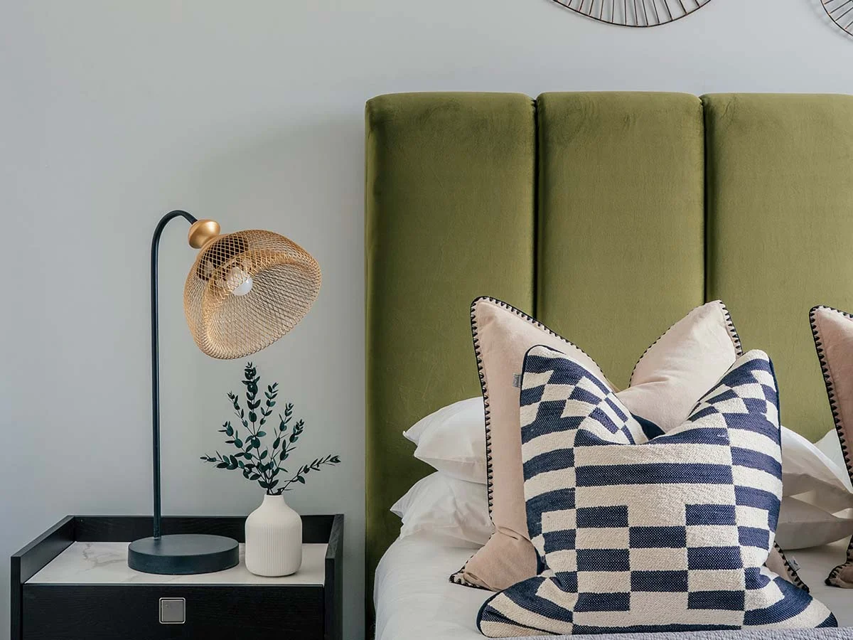 A modern bedroom interior styled by InStyle Direct showcasing a vertical-panelled olive green headboard, monochrome geometric patterned pillows, and a contemporary gold mesh dome table lamp in Fulham
