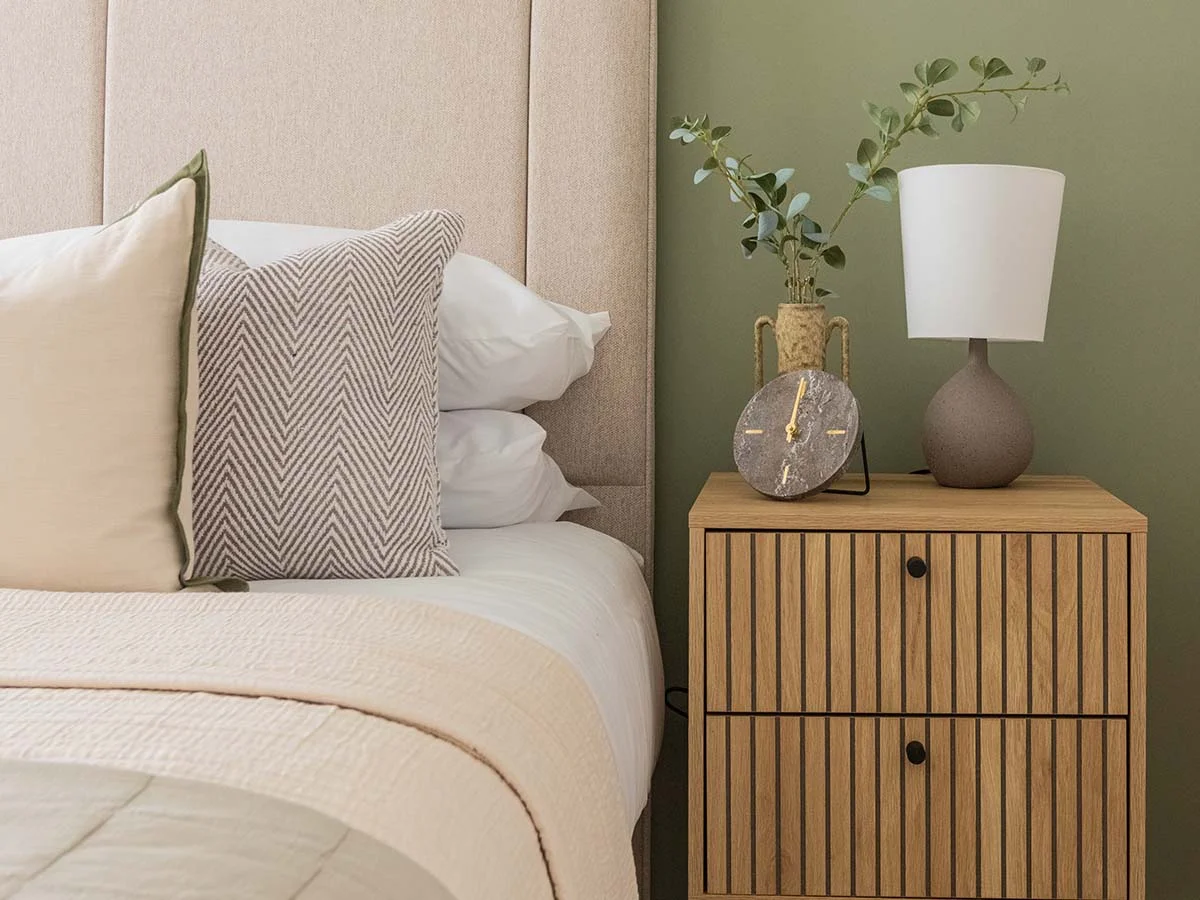 Close-up of a slatted timber nightstand styled by InStyle Direct at Regents View, featuring a round marble desk clock, a ceramic lamp against a sage green wall, and chevron-patterned pillows