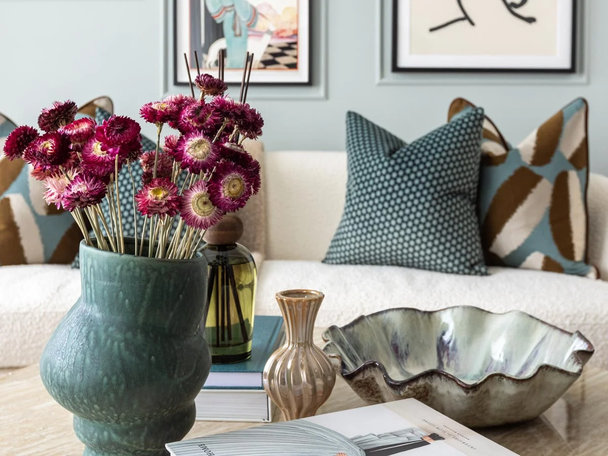 Close-up styling of a coffee table featuring a green ceramic vase filled with vibrant pink dried flowers, decorative bowls, and textured scatter cushions on a white sofa