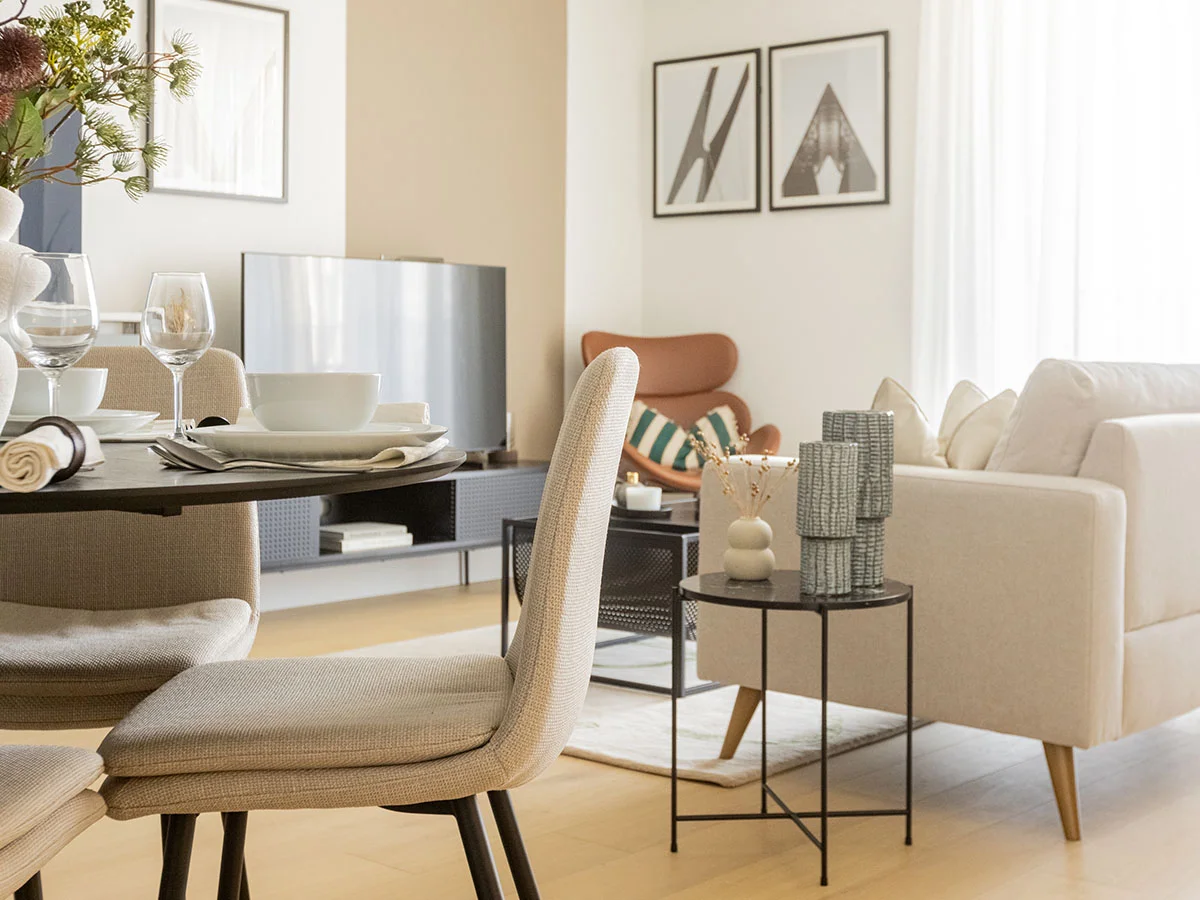 An open-plan apartment space professionally staged by InStyle Direct, capturing the transition between an intimate dining setting with beige chairs and a bright, sunlit reception lounge.