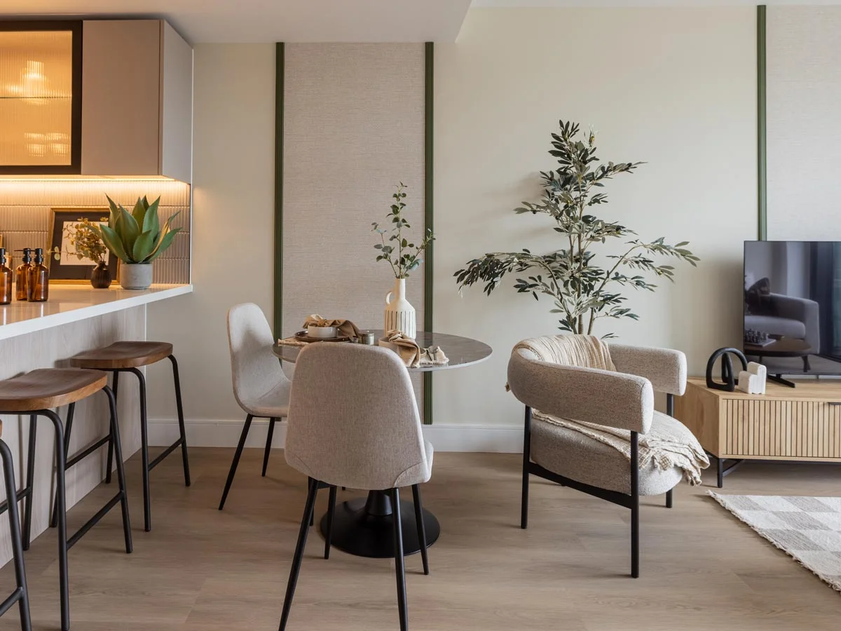 A modern open-plan apartment interior styled by InStyle Direct, featuring a breakfast bar with curved walnut stools, a round marble-topped dining table with beige chairs, and a contemporary grey accent chair next to a potted olive tree