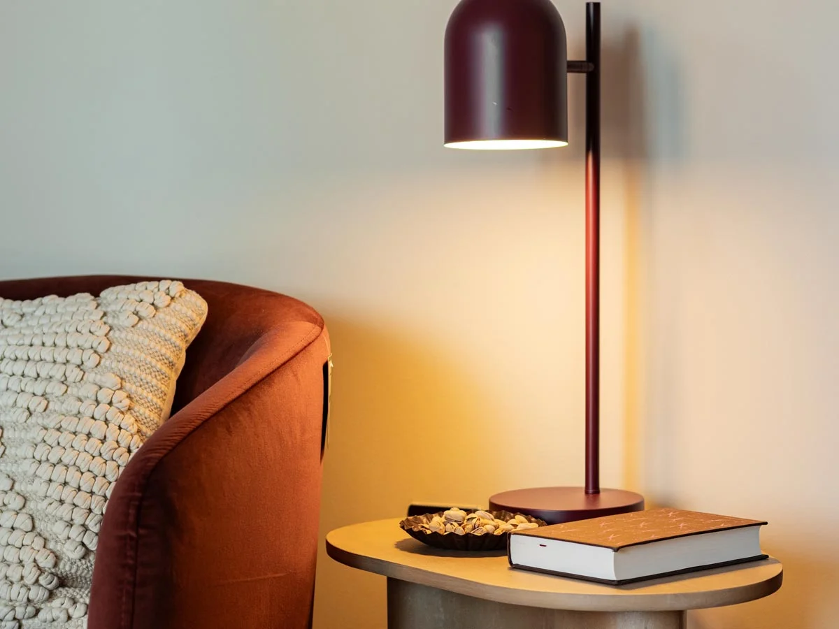 Close-up of a modern light oak side table styled by InStyle Direct featuring a minimalist matte burgundy dome table lamp, a textured rust armchair, and a decorative tray with snacks