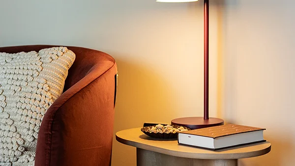 Close-up of a modern light oak side table styled by InStyle Direct featuring a minimalist matte burgundy dome table lamp, a textured rust armchair, and a decorative tray with snacks