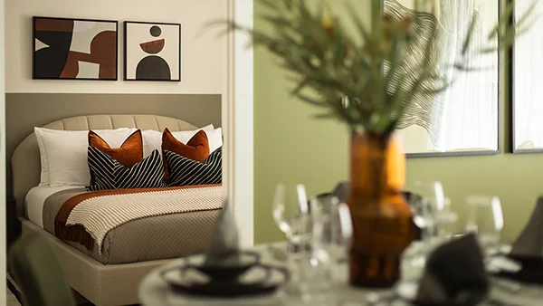 An apartment staging perspective by InStyle Direct at the Verdica show flats, looking across a dressed dining table toward a serene bedroom with a fluted headboard, rust velvet pillows, and abstract wall prints