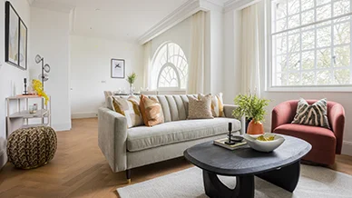 A premium open-plan apartment reception lounge styled by InStyle Direct at Moda Holland Park, featuring a fluted grey sofa, a terracotta accent chair, a dark wood coffee table, and large bright windows