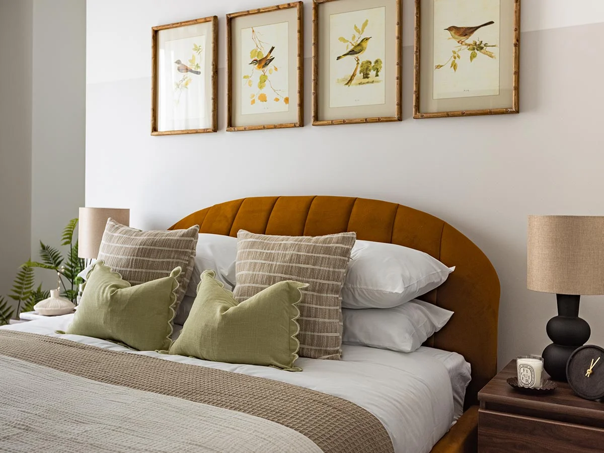 Elegant bedroom interior featuring a curved mustard yellow tufted headboard, framed botanical wall art, striped cushions, and bedside table lamps at Regent's View