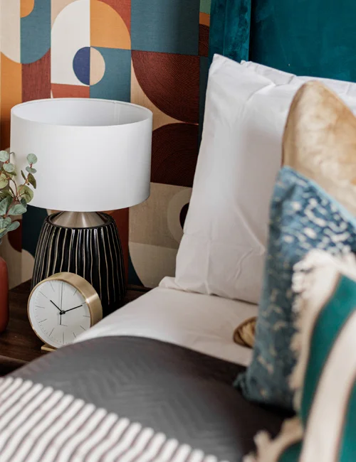 A retro bedroom detail styled by InStyle Direct featuring bold multi-coloured geometric wallpaper, a plush teal headboard, and a dark wood nightstand with a white table lamp and gold clock in Islington