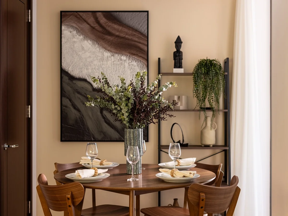 A round walnut dining table setting styled by InStyle Direct at West End Gate, featuring mid-century wooden chairs, fine stemware, a tall eucalyptus vase, and a open shelving unit