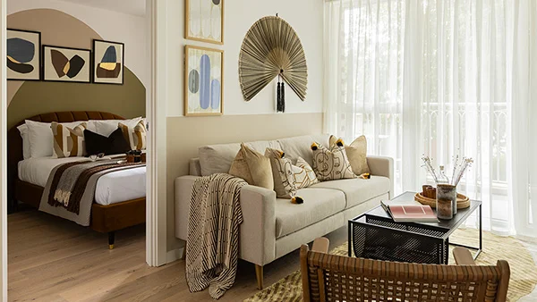 A stylish show flat interior by InStyle Direct at Verdica, showing a bright living room with woven fan wall art, a neutral sofa, and an open doorway through to a staged bedroom with an ochre velvet headboard