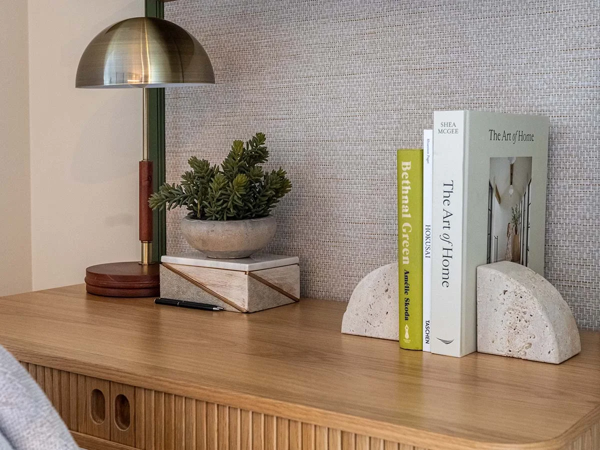 A detailed view of a slatted wood desk workspace styled by InStyle Direct at Regents View, showcasing travertine stone bookends, a dome desk lamp, and a small potted succulent